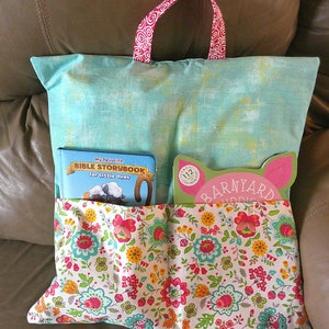 May include: A turquoise pillow with two pockets, one holding a book titled "My Favorite Bible Storybook for Little Ones" and the other holding a book titled "Barnyard Hoot". The pillow has a floral print with pink, yellow, and green flowers on a white background.