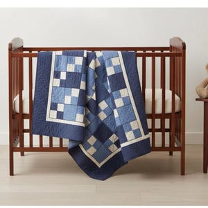 May include: A dark brown wooden crib with a blue and white patchwork quilt draped over the side. A teddy bear sits on a small side table. The quilt features a square pattern in shades of blue and white.