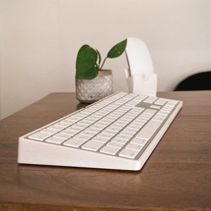 Könnte beinhalten: Eine weiße drahtlose Tastatur mit silbernem Rand liegt auf einem Holztisch. Im Hintergrund sind eine kleine Topfpflanze und eine weiße drahtlose Maus zu sehen.