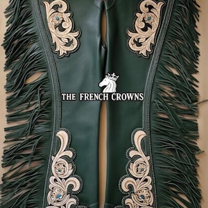 May include: Dark green leather chaps with intricate cream-colored floral designs and turquoise accents. The chaps feature long fringe along the sides and are embellished with silver-toned studs. The text "THE FRENCH CROWNS" is visible.