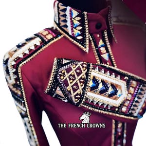 Könnte beinhalten: Ein weinrotes Reitshirt mit aufwendigen Verzierungen. Kragen und Manschetten weisen aufwendige Perlenarbeiten in Gold, Schwarz und Juwelfarben auf. Der Text "THE FRENCH CROWNS" ist unten aufgedruckt.