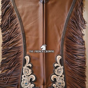 May include: Brown leather chaps featuring long fringe along the outer edges, a dark brown waistband with silver-toned studs, and light-colored floral tooling at the bottom. The text THE FRENCH CROWNS with a horse logo is printed in the center.