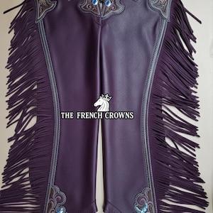May include: Purple leather chaps with decorative silver and brown filigree, blue gemstones, and long fringe. The text "THE FRENCH CROWNS" is printed on the chaps. These chaps are designed for riding and equestrian activities.