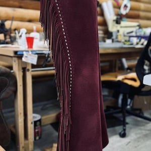 Op de afbeelding: Bordeaux leren chaps met decoratieve bloemmotieven en franjes. De chaps hebben een gedetailleerde leren tailleband en zijn versierd met zilveren studs. Een kledingstuk in westernstijl.