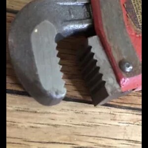 May include: Close-up of a red and silver adjustable wrench with a serrated jaw. The wrench is resting on a wooden surface.