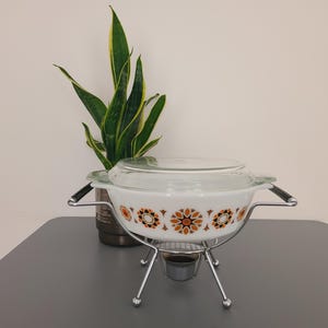 May include: A retro white casserole dish with a glass lid, decorated with an orange and brown floral design. It is on a chrome stand with black handles. A potted plant with green and yellow leaves is in the background.