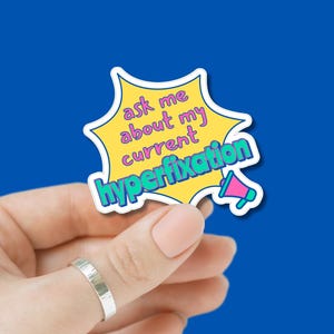 Op de afbeelding: Een sticker met een geel sterrenburst-ontwerp en de tekst "ask me about my current hyperfixation" in blauw en roze. De sticker wordt tegen een blauwe achtergrond gehouden.