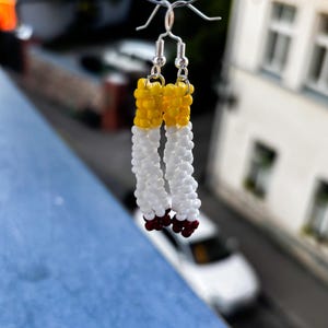 Op de afbeelding: Handgemaakte kralen oorbellen in de vorm van sigaretten. Elke oorbel heeft een gele punt, een witte body en een roodbruin uiteinde. De oorbellen hangen aan zilverkleurige haken.