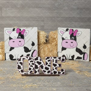 Peut inclure: Lettres en bois "UN" décorées d'un imprimé vache noir et blanc. Les lettres sont posées sur une base brune. Deux panneaux à thème vache sont en arrière-plan, chacun représentant une vache noire et blanche avec un nœud rose.