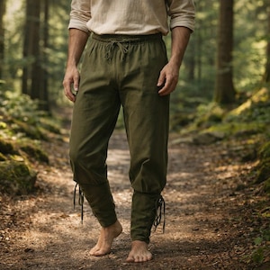 May include: Olive green drawstring pants with ankle ties. The pants are made of a soft fabric and are worn with a light-colored long-sleeve shirt. The person is standing on a dirt path in a forest.