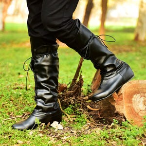 K&ouml;nnte beinhalten: Schwarze kniehohe Stiefel mit Schn&uuml;rsenkeln. Die Stiefel haben eine glatte, gl&auml;nzende Oberfl&auml;che und einen kleinen Absatz. Die Schn&uuml;rsenkel verlaufen &uuml;ber die Vorderseite der Stiefel. Die Stiefel werden mit einer schwarzen Hose getragen. Die Stiefel stehen auf einem Baumstumpf.