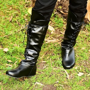 K&ouml;nnte beinhalten: Schwarze kniehohe Stiefel mit Schn&uuml;rdesign an der Seite. Die Stiefel haben eine glatte, gl&auml;nzende Oberfl&auml;che und werden &uuml;ber einer schwarzen Hose getragen. Die Stiefel werden vor einem Hintergrund aus gr&uuml;nem Gras und Laub gezeigt.