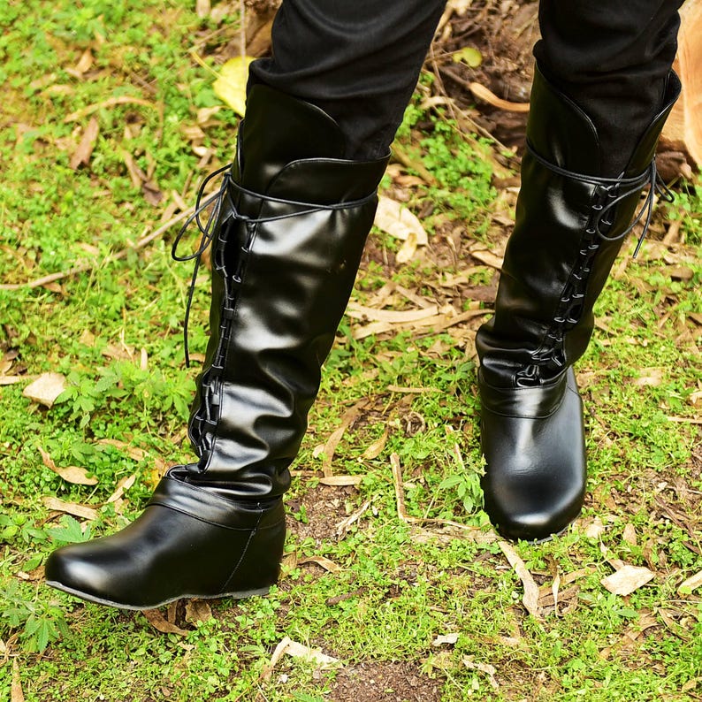 K&ouml;nnte beinhalten: Schwarze kniehohe Stiefel mit Schn&uuml;rdesign an der Seite. Die Stiefel haben eine glatte, gl&auml;nzende Oberfl&auml;che und werden &uuml;ber einer schwarzen Hose getragen. Die Stiefel werden vor einem Hintergrund aus gr&uuml;nem Gras und Laub gezeigt.