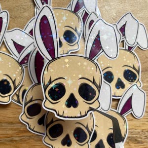 May include: A collection of stickers featuring skull designs with bunny ears. The skulls have black eye sockets and are decorated with sparkling stars. The bunny ears are white with a purple interior.