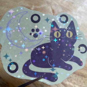 May include: A holographic sticker depicting a black cat with yellow eyes, stars, a crescent moon, and paw prints. The sticker has a light green background with a glittery, iridescent finish, and black circles.
