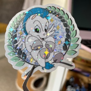 Könnte beinhalten: Ein holografischer Aufkleber mit einem weißen Cartoon-Pegasus mit blauen Flügeln und einer blauen Mähne. Der Pegasus ist von einem grünen Lorbeerkranz und einem glitzernden Hintergrund umgeben.