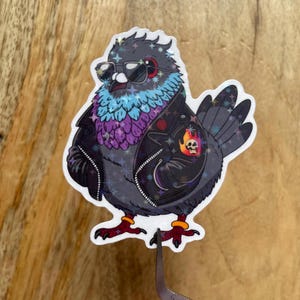 Può includere: Un adesivo con un cartone animato di un piccione che indossa occhiali da sole e una giacca di pelle nera con una toppa di teschio e fiamme. Il piccione ha un bagliore iridescente blu e viola.