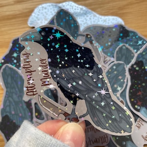 Op de afbeelding: Een zwarte raaf sticker met een mes in zijn klauwen. De raaf is bedekt met iriserende glitter en heeft een sterrenhemel achtergrond. De sticker zegt "Na een moordpoging".