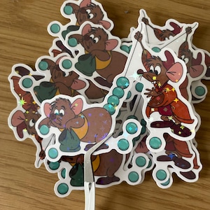 Op de afbeelding: Een stapel stickers met een cartoonmuisfiguur in verschillende poses. De muis draagt ​​een bruin jasje en houdt een groene tas vast. De stickers zijn iriserend en hebben een glinsterende afwerking.