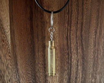 Könnte beinhalten: Ein goldfarbener Patronenhülsen-Anhänger an einer schwarzen Kordelkette. Der Anhänger ist mit einer silbernen Kette an der Kordel befestigt.