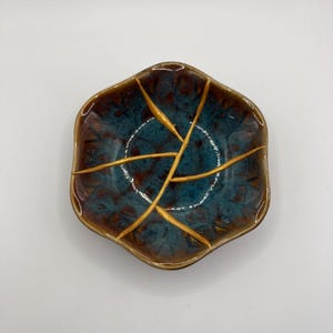 Puede incluir: Un pequeño plato hexagonal de cerámica con un esmalte azul y marrón. El plato tiene un acabado craquelado con detalles dorados.