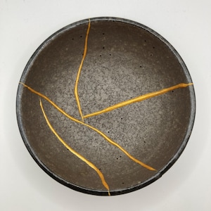 Könnte beinhalten: Dunkelgraue Keramikschale mit schwarzem Rand, versehen mit goldfarbenen Kintsugi-Reparaturen. Die Schale hat eine strukturierte Oberfläche und sichtbare Unvollkommenheiten. Die goldenen Linien heben die Risse hervor.