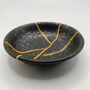 May include: A small, round, black ceramic dish with a speckled pattern. The dish features gold-coloured lines that appear to mend cracks, showcasing the Japanese art of Kintsugi. The dish is set against a white background.