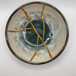 Op de afbeelding: Een witte keramische kom met een blauwe en grijze swirl-patroon. De kom heeft gouden scheuren over het oppervlak.