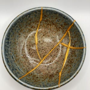 Könnte beinhalten: Eine blaue Keramikschüssel mit gesprenkeltem Glase und goldenen Akzenten. Die Schüssel hat eine runde Form und einen leicht ausgestellten Rand.