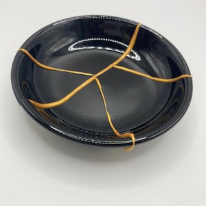 Op de afbeelding: Een zwarte, ovale keramische kom met goudkleurige kintsugi reparatielijnen. De kom heeft een glanzende afwerking en staat tegen een witte achtergrond. De gouden lijnen benadrukken de scheuren en tonen de Japanse kunst van het repareren van gebroken aardewerk.