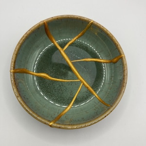 May include: A round, teal ceramic bowl featuring a gold kintsugi repair design. The bowl has a speckled texture and a brown rim. The gold lines provide a striking contrast against the teal background, showcasing the Japanese art of repairing broken pottery.
