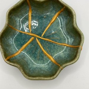 Può includere: Una ciotola in ceramica verde e turchese con una forma organica unica. La ciotola presenta crepe visibili, riparate con accenti color oro, mostrando l'arte giapponese del Kintsugi. Il bordo della ciotola ha un design ondulato.
