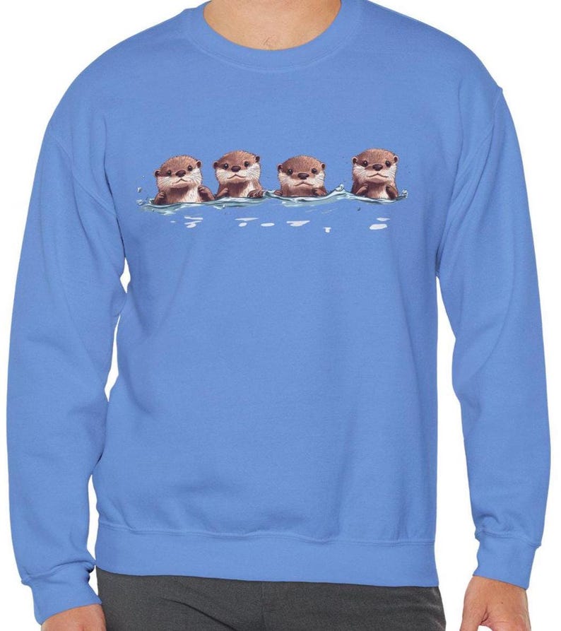 Op de afbeelding: Een blauw sweatshirt met een afbeelding van vier otters die in een rij zwemmen. De otters zijn bruin en hebben witte buiken. De afbeelding is gecentreerd op het sweatshirt.