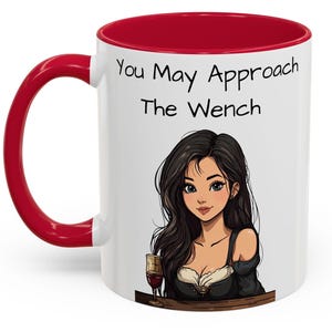 May include: White ceramic mug with a red rim and handle. The mug has a cartoon image of a woman with long black hair and a glass of wine. The text "You May Approach The Wench" is printed on the mug.