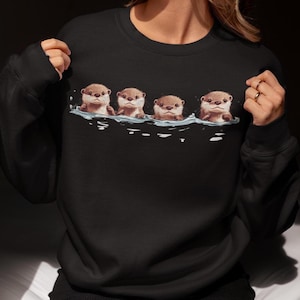 Op de afbeelding: Zwart sweatshirt met een afbeelding van vier otters die in een rij zwemmen. De otters zijn bruin en hebben witte gezichten.