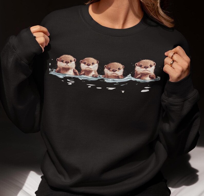 Op de afbeelding: Zwart sweatshirt met een afbeelding van vier otters die in een rij zwemmen. De otters zijn bruin en hebben witte gezichten.