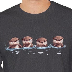 Op de afbeelding: Een donkergrijs sweatshirt met een afbeelding van vier otters die in een rij zwemmen. De otters zijn bruin en hebben schattige gezichten.