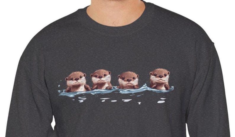 Op de afbeelding: Een donkergrijs sweatshirt met een afbeelding van vier otters die in een rij zwemmen. De otters zijn bruin en hebben schattige gezichten.