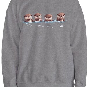 Op de afbeelding: Een grijs sweatshirt met een afbeelding van vier otters die in een rij zwemmen. De otters zijn bruin en hebben schattige gezichten.