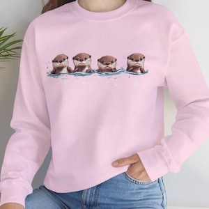 Op de afbeelding: Een roze sweatshirt met een afbeelding van vier otters die in een rij zwemmen. De otters zijn bruin en hebben een witte buik.