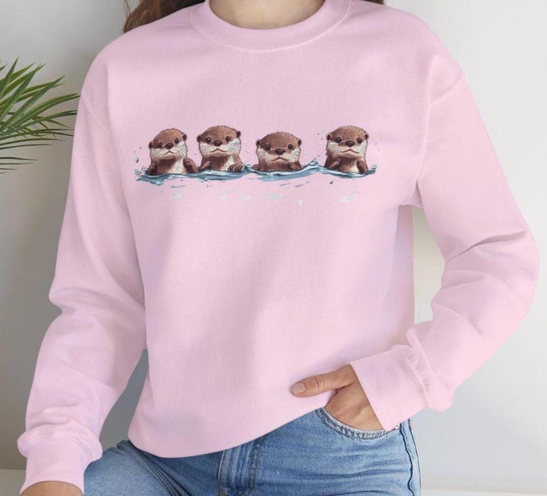 Op de afbeelding: Een roze sweatshirt met een afbeelding van vier otters die in een rij zwemmen. De otters zijn bruin en hebben een witte buik.
