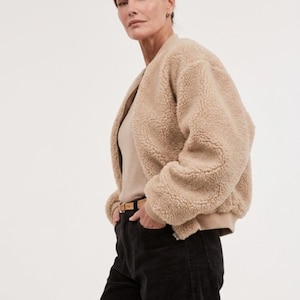 Puede incluir: Chaqueta de osito de peluche beige con un corte relajado. La chaqueta tiene un diseño sin cuello y un dobladillo y puños acanalados. La modelo lleva pantalones de pana negros y un cinturón marrón. La chaqueta está hecha de un material suave y afelpado.