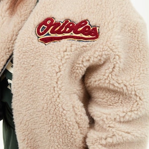 Könnte beinhalten: Beige, flauschige Bomberjacke mit schwarzem Kragen und Reißverschluss. Die Jacke hat einen Aufnäher mit dem Wort "Orioles" in roter und weißer Schrift. Die Jacke ist aus einem weichen, strukturierten Material.