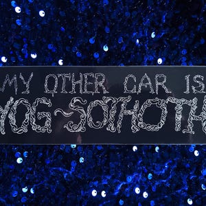 May include: Black rectangular bumper sticker with the text "MY OTHER CAR IS YOG-SOTHOTH" in white, stylized lettering. The sticker is set against a background of blue sequins.