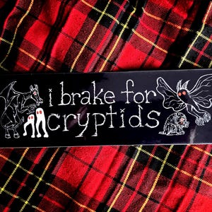 May include: Black rectangular sticker with white text and illustrations. The text reads "i brake for cryptids." Illustrations include a winged creature, a moth-like figure, a small furry beast, and two ghost-like figures.