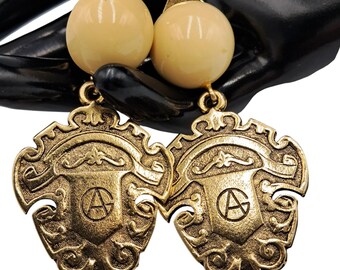 May include: Vintage gold-tone clip-on earrings. Each earring features a cream-colored ball and a shield-shaped pendant with an embossed design and the letter "A" inside a circle. The earrings have a classic, ornate style.