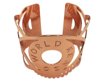 Könnte beinhalten: Roségoldenes Armreif-Armband mit den Worten "KENZO WORLD" auf einem kreisförmigen Mittelstück eingraviert. Das offene Design weist dekorative Ausschnitte und eine polierte Oberfläche auf, geeignet zum Tragen am Handgelenk.