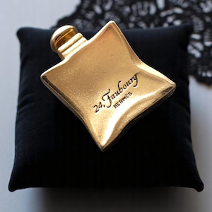 May include: A gold-colored perfume bottle with a unique, geometric shape, resting on a black velvet pillow. The bottle has a small, cylindrical cap and the text "24. Faubourg HERMÈS" is inscribed on the front.