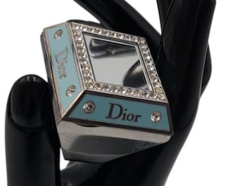 Puede incluir: Un anillo de tono plateado con una cara rectangular que presenta un panel de espejo central rodeado de cristales transparentes. Los lados del anillo están acentuados con esmalte azul claro y el texto Dior, con cristales incrustados.