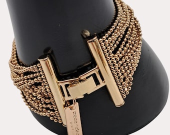 May include: Gold-toned bracelet featuring multiple strands of small beads. The bracelet has a rectangular clasp with a small tag attached. The bracelet is displayed on a black form.